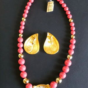 Pu&ograve; includere: Una collana di perline rosa corallo con un ciondolo dorato e orecchini abbinati. Il ciondolo ha la forma di una foglia e una superficie testurizzata. Anche gli orecchini hanno la forma di una foglia e una texture simile. La collana ha una chiusura a moschettone.