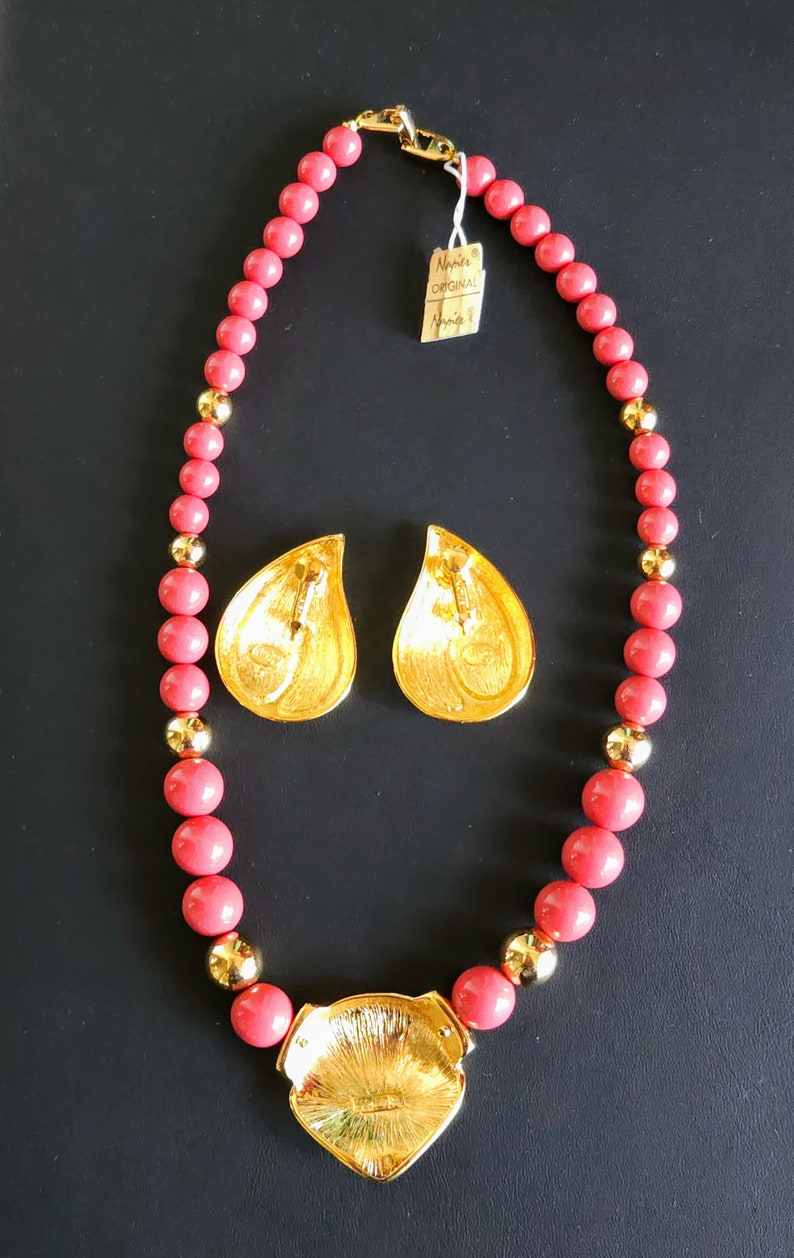 Pu&ograve; includere: Una collana di perline rosa corallo con un ciondolo dorato e orecchini abbinati. Il ciondolo ha la forma di una foglia e una superficie testurizzata. Anche gli orecchini hanno la forma di una foglia e una texture simile. La collana ha una chiusura a moschettone.