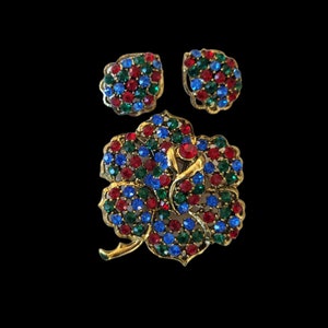 Pode incluir: Um conjunto de broche e brincos em tom dourado com um design floral. As joias s&atilde;o adornadas com strass vermelhos, azuis e verdes.