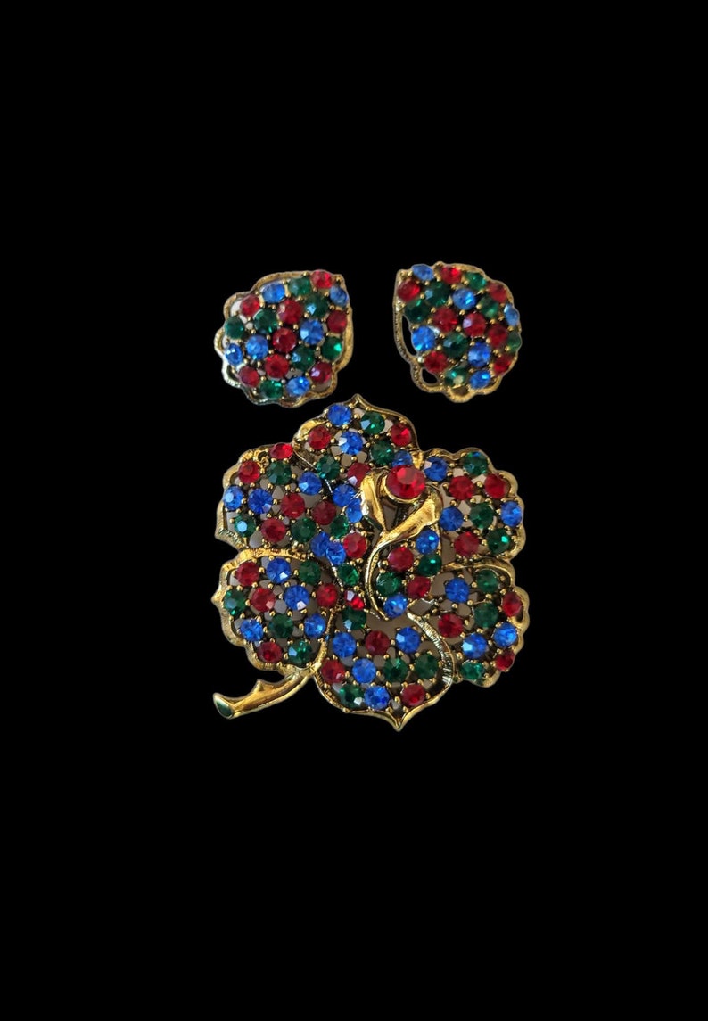 Pode incluir: Um conjunto de broche e brincos em tom dourado com um design floral. As joias s&atilde;o adornadas com strass vermelhos, azuis e verdes.