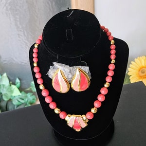 Pu&ograve; includere: Una collana di perline rosa e oro con un paio di orecchini abbinati. La collana ha un grande ciondolo al centro, a forma di cuore con un design geometrico. Gli orecchini hanno la forma di una goccia.