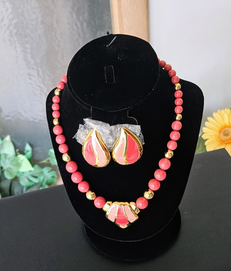 Pu&ograve; includere: Una collana di perline rosa e oro con un paio di orecchini abbinati. La collana ha un grande ciondolo al centro, a forma di cuore con un design geometrico. Gli orecchini hanno la forma di una goccia.