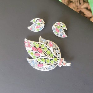 Pu&ograve; includere: Un set di spilla e orecchini in tono argento con un design floreale. La spilla presenta una grande forma di foglia con strass rosa, verdi e blu. Gli orecchini sono pi&ugrave; piccoli e presentano una forma di foglia simile con strass rosa e blu.