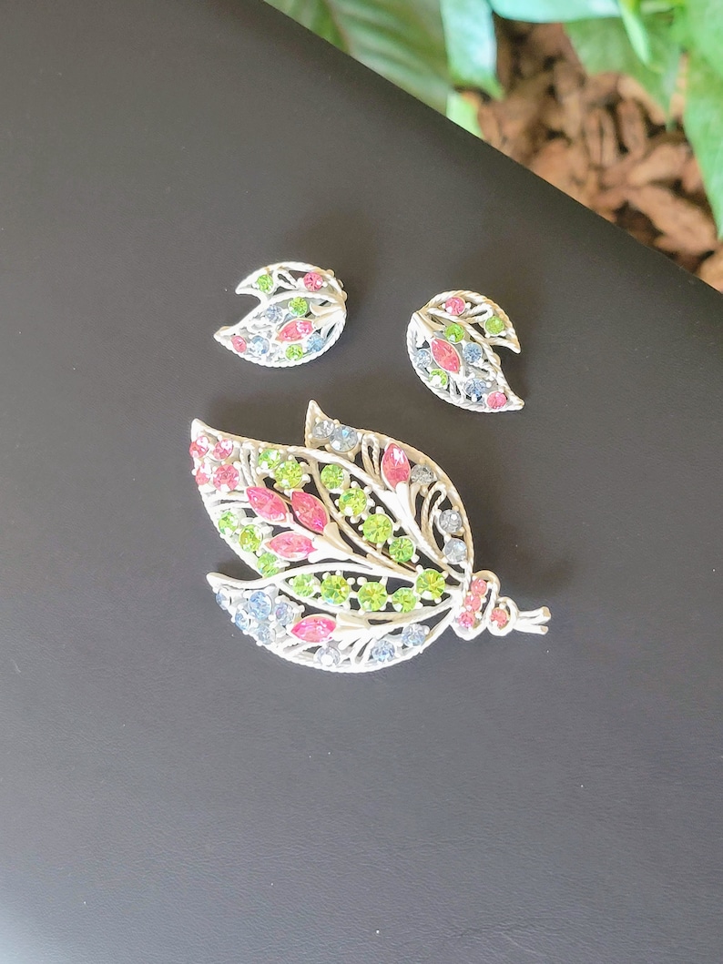 Pu&ograve; includere: Un set di spilla e orecchini in tono argento con un design floreale. La spilla presenta una grande forma di foglia con strass rosa, verdi e blu. Gli orecchini sono pi&ugrave; piccoli e presentano una forma di foglia simile con strass rosa e blu.