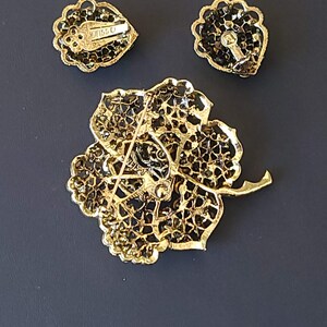 Pode incluir: Um conjunto de broche e brincos dourados com um design floral e strass pretos. O broche tem um fecho de pino e os brincos t&ecirc;m fechos de press&atilde;o. O broche tem o texto "Weiss" na parte de tr&aacute;s.