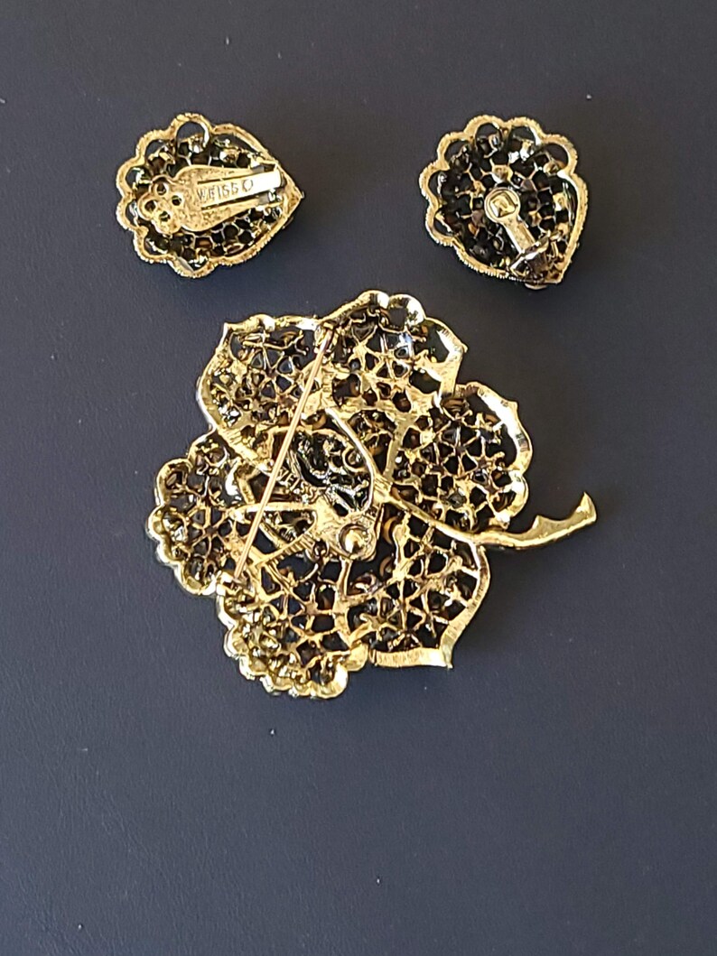 Pode incluir: Um conjunto de broche e brincos dourados com um design floral e strass pretos. O broche tem um fecho de pino e os brincos t&ecirc;m fechos de press&atilde;o. O broche tem o texto "Weiss" na parte de tr&aacute;s.