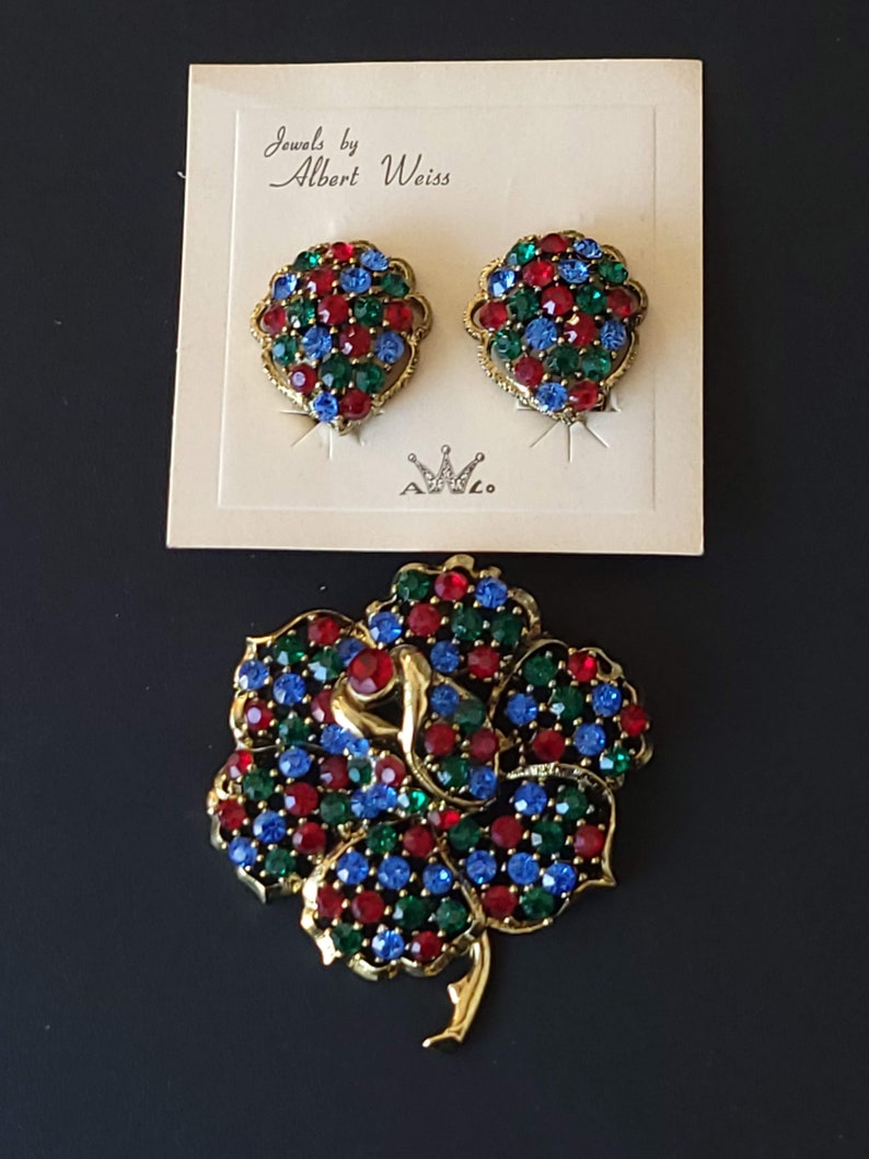 Pode incluir: Um conjunto de joias vintage da Albert Weiss, incluindo um broche e brincos. O broche &eacute; uma flor dourada com pedras de strass vermelhas, azuis e verdes. Os brincos s&atilde;o cachos correspondentes de pedras de strass vermelhas, azuis e verdes.