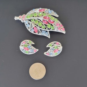 Pu&ograve; includere: Un set di gioielli vintage composto da una grande spilla a forma di foglia e due orecchini abbinati. I gioielli sono realizzati in metallo color argento e sono adornati con strass colorati in tonalit&agrave; rosa, verde e blu.