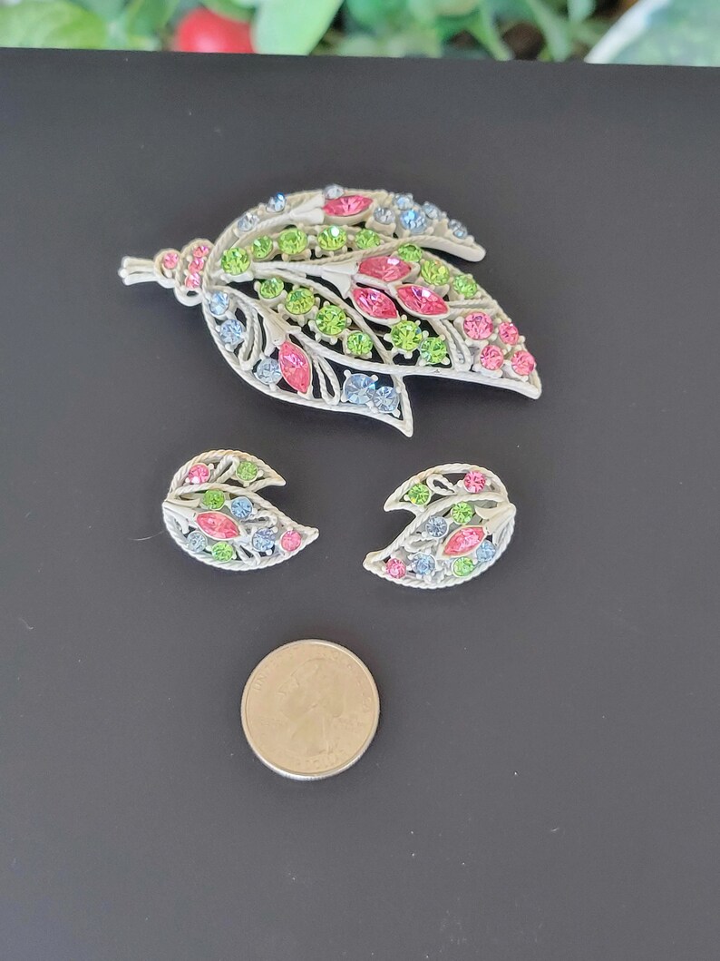 Pu&ograve; includere: Un set di gioielli vintage composto da una grande spilla a forma di foglia e due orecchini abbinati. I gioielli sono realizzati in metallo color argento e sono adornati con strass colorati in tonalit&agrave; rosa, verde e blu.