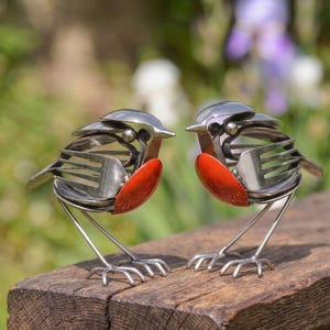 May include: Two bird sculptures crafted from repurposed stainless steel cutlery, including forks and spoons. Each bird features a distinct bright orange painted chest, metallic silver wings made from fork tines, and thin metal wire legs standing upright.