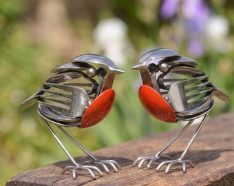 May include: Two bird sculptures crafted from repurposed stainless steel cutlery, including forks and spoons. Each bird features a distinct bright orange painted chest, metallic silver wings made from fork tines, and thin metal wire legs standing upright.
