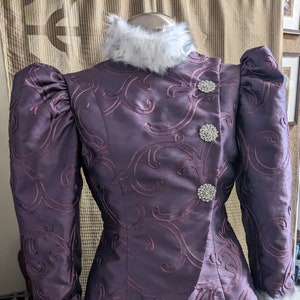 May include: A purple dress with a white fur collar and cuffs. The dress has a fitted bodice with a decorative pattern and three large, clear rhinestone buttons. The skirt is full and flowing.