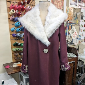 May include: A long, burgundy coat with a large white fur collar. The coat has a single button closure and beaded embellishments on the cuffs.