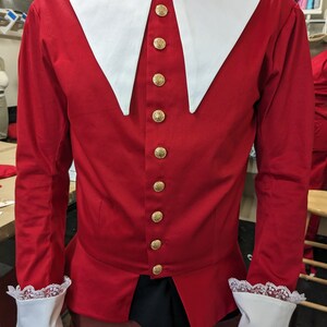 May include: A red long-sleeved shirt with a white collar and gold buttons. The shirt has a pointed collar and lace trim on the cuffs.