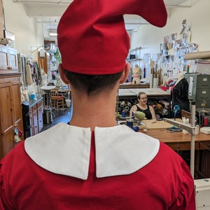 May include: A person wearing a red costume with a white collar and a red pointed hat. The costume is reminiscent of a traditional jester or elf outfit.