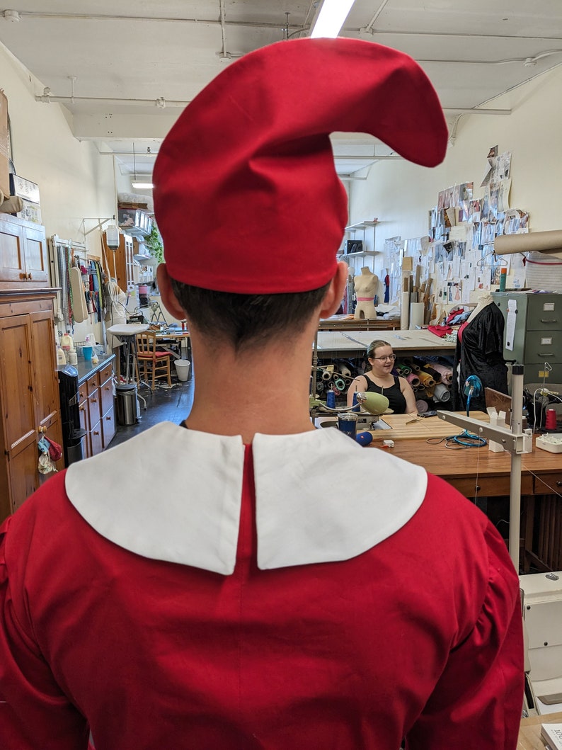 May include: A person wearing a red costume with a white collar and a red pointed hat. The costume is reminiscent of a traditional jester or elf outfit.
