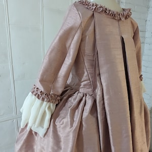 Puede incluir: Un vestido de color rosa polvoriento con falda fruncida y mangas largas. Las mangas tienen volantes blancos en los puños. El vestido tiene una abertura en la espalda con una costura central.