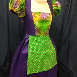 May include: A purple dress with a green and yellow floral print bodice and a green apron with gold fleur-de-lis print. The dress has puffed sleeves and a white ruffle collar.