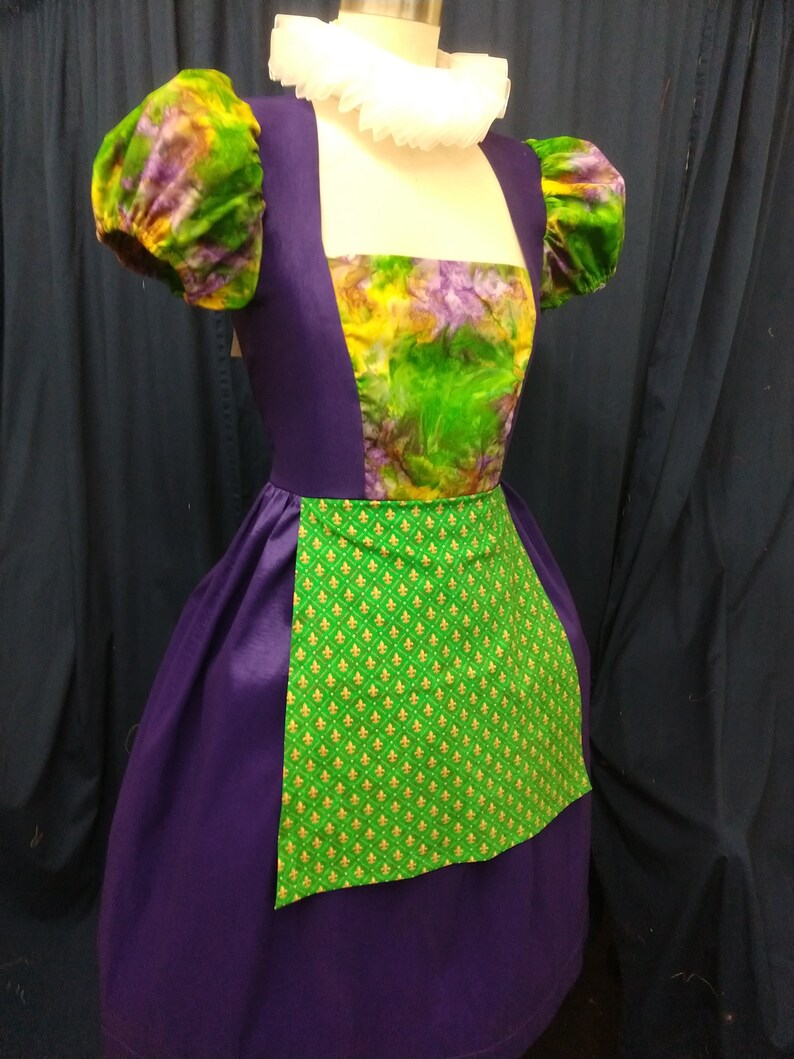 May include: A purple dress with a green and yellow floral print bodice and a green apron with gold fleur-de-lis print. The dress has puffed sleeves and a white ruffle collar.