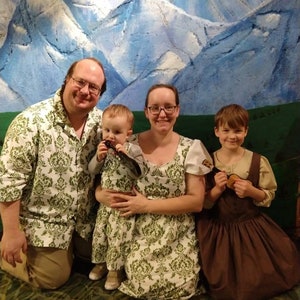 May include: A family of four poses in front of a painted mountain backdrop. The adults and baby wear matching white and green patterned outfits. The child on the right wears a brown dress with a tan top.