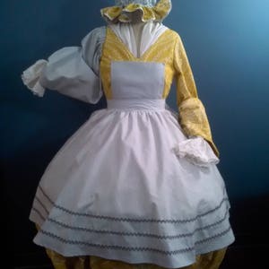 Puede incluir: Un vestido blanco con un corpiño amarillo y un delantal blanco. El vestido tiene un cuello y puños con volantes. El vestido lo lleva un maniquí con un sombrero azul y amarillo.