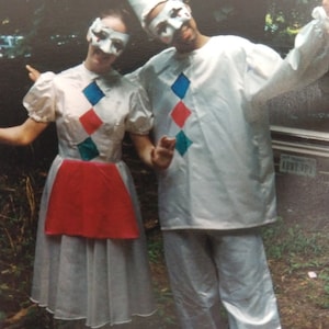 May include: Two people wearing white costumes with colorful diamond patterns and masks. The person on the left is wearing a red skirt and a white dress. The person on the right is wearing a white shirt and pants.