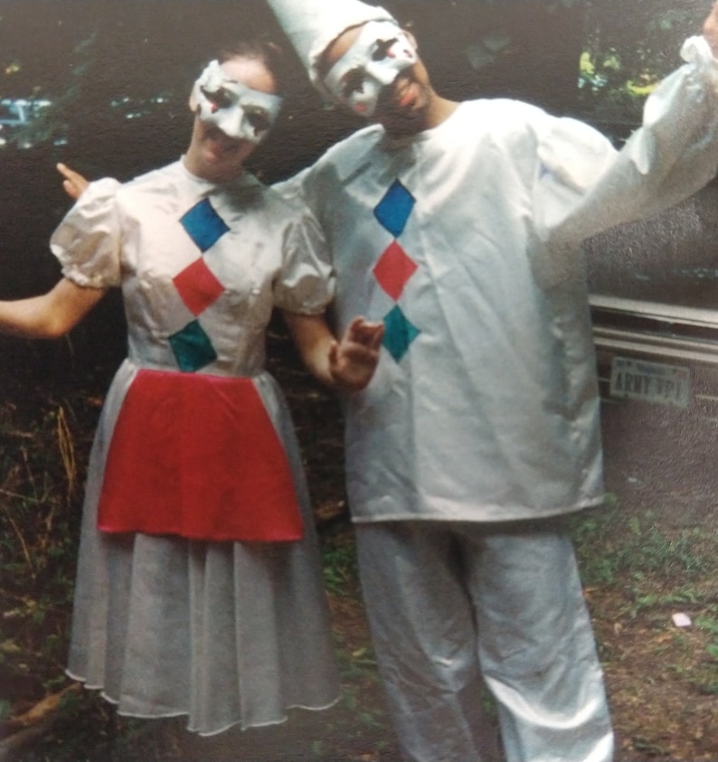 May include: Two people wearing white costumes with colorful diamond patterns and masks. The person on the left is wearing a red skirt and a white dress. The person on the right is wearing a white shirt and pants.