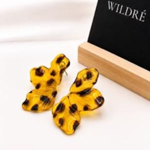 May include: A pair of translucent amber-colored earrings with a dark brown leopard print pattern. The earrings have an irregular, organic, wavy shape and are displayed next to a black sign with the white text WILDRE on a wooden base.