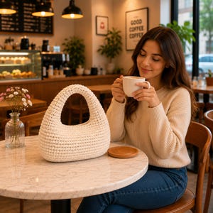 May include: A cream-colored, crescent-shaped handbag crafted from a thick, textured knit material. The bag features a single integrated handle that forms a continuous arch with the body, showcasing a dense, uniform stitch pattern throughout.