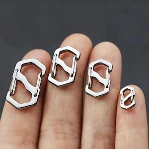 May include: Four silver metal carabiner clips of different sizes. The clips have a distinctive design with a central bar and a hexagonal base. The smallest clip is on a fingertip. The clips are made of metal.