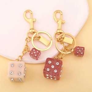 May include: Two gold-colored keychains with dice charms. One keychain has a light pink dice charm, and the other has a brown dice charm. Both dice are embellished with clear rhinestones. The keychains have a gold clasp and a small gold tag.