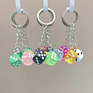 May include: A collection of keychains with colorful dice charms. Each keychain features a silver ring, a chain, and a translucent die in various colors, including green, pink, black, and yellow. The dice have white dot markings.