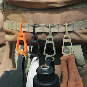 Può includere: Quattro clip utilitarie in plastica arancione, nera, verde oliva e beige sono attaccate a uno zaino. Le clip tengono un cappello nero, un panno bianco, una bottiglia d'acqua nera e un calzino marrone. Le clip sono progettate per l'attrezzatura da esterno.