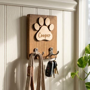 Könnte beinhalten: Ein hölzerner Schlüsselhalter mit einem Pfotenabdruck-Design und dem eingravierten Namen "Cooper". Er hat zwei Metallhaken für Schlüssel und eine Leine. Der Schlüsselhalter ist an einer weißen Wand mit Paneelen befestigt, mit einer Pflanze im Hintergrund.