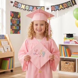 Peut inclure: Une jeune fille porte une robe et un chapeau de graduation roses, tenant un chapeau assorti brodé du nom "Lily". L'arrière-plan comprend des ballons colorés, une bannière avec "Congrats Grads!" et une étagère remplie de livres.