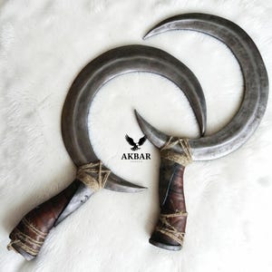 May include: Two curved, crescent-shaped blades with dark metal finishes. The handles are wrapped in brown leather and twine. The image includes the text "AKBAR FORGE" with a stylized eagle emblem.