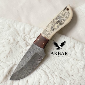 Puede incluir: Un cuchillo artesanal con una hoja de acero estampada y un mango de madera. El mango presenta un diseño grabado y está conectado a una sección crema con un diseño de pájaro grabado. El cuchillo está etiquetado como "AKBAR FORGED".