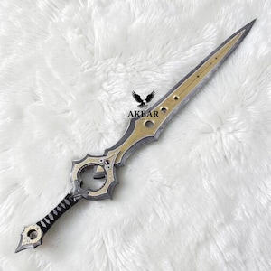 May include: A decorative dagger featuring a pointed blade with a central gold-coloured inlay, a circular guard with a small purple gem, and a black handle. The blade is engraved with the text AKBAR and a graphic of an eagle with wings spread.