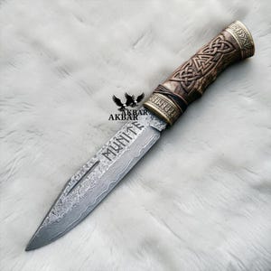 Puede incluir: Un cuchillo decorativo con una hoja estampada y un mango de madera con diseños de nudos celtas. La hoja tiene grabada la palabra "MUNITE". El mango está acentuado con metal color latón. El cuchillo se exhibe sobre una superficie blanca texturizada.