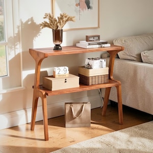 May include: A two-tiered wooden side table in a warm, reddish-brown hue. The table has a curved design and displays decorative items such as a vase with dried flowers, books, a clock, and a woven basket. A paper bag is placed below.
