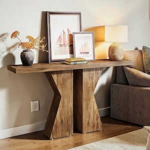May include: A wooden console table with a distinctive geometric leg design. The table features framed sailboat art, a lamp, a vase of flowers, and books. The warm wood tones and neutral decor create a cosy, inviting atmosphere.