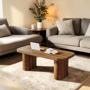 May include: A rectangular coffee table with a dark wood grain finish and rounded corners, supported by two thick, curved pillar-style legs. A white laptop and an open book are placed on the flat surface of the table, which sits on a rug.