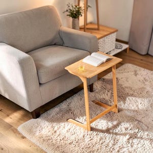 May include: A light-coloured wooden side table with a rectangular top and a unique angled leg design. An open book and a glass of liquid sit on the table. The table is next to a grey armchair on a fluffy rug.