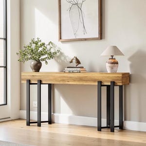 May include: A rectangular console table with a light brown wood grain top and two sets of black metal legs. The table surface holds a green potted plant in a dark vase, a small bronze horse sculpture, stacked books, and a table lamp with a beige shade.