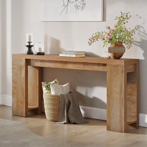 May include: A light brown wooden console table with a rectangular top and two thick legs. A woven basket with pillows and a blanket sits below the table. A vase with flowers, candles, and books are on top.
