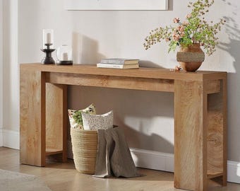May include: A light brown wooden console table with a rectangular top and two thick legs. A woven basket with pillows and a blanket sits below the table. A vase with flowers, candles, and books are on top.