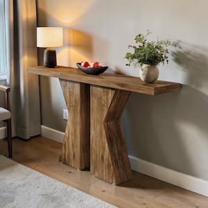 May include: A wooden console table with a natural finish. The table has a long, rectangular top supported by two angled, X-shaped legs. A lamp, a bowl of fruit, and a vase of greenery are displayed on the table.
