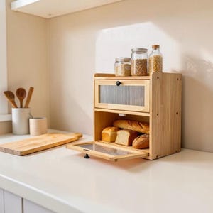May include: A two-tier wooden bread box with a light natural finish, featuring a drop-down door with a ribbed glass panel. The interior holds various loaves of bread, while the top surface displays three clear glass jars filled with grains.