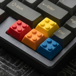 May include: A close-up of a mechanical keyboard with custom keycaps shaped like colorful toy building blocks. The keycaps are red, orange, yellow, and blue, contrasting with the dark gray keys and keyboard frame.