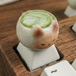 May include: A custom mechanical keyboard keycap shaped like a small ceramic cat mug filled with green matcha latte art. The keycap is white with brown ear details and sits on a wooden keyboard frame alongside other white keys with black text.